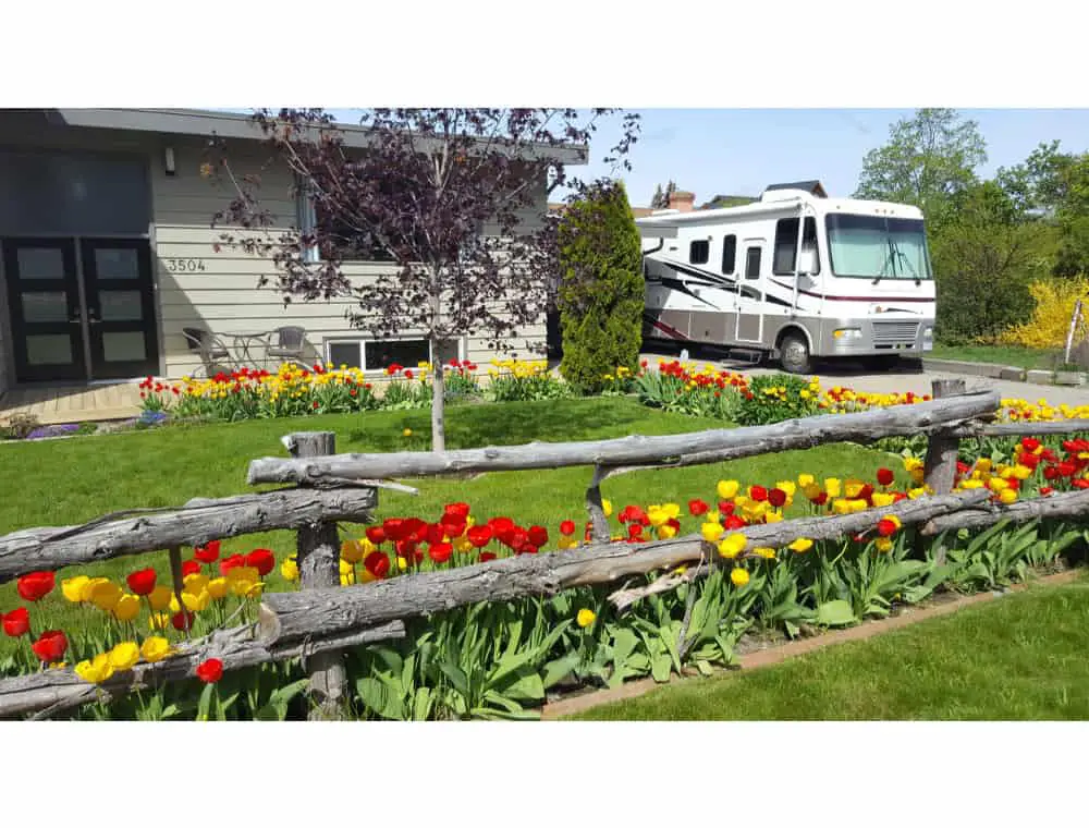 A big RV parked on a driveway in a beautiful garden full of yellow and red flowers of a ravishing house