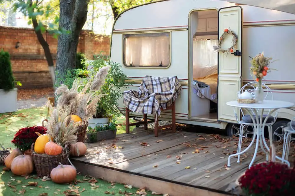 An RV parked in a garden of a house with a proper set up of chairs, a big platform, pumpkins, and greenery